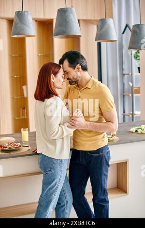 A bearded man and a redhead woman are happily dancing together in a sunlit modern kitchen. Stock Photo