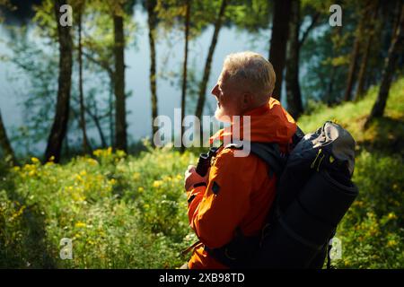 High angle view of senior male tourist wearing orange jacket with gray backpack looking at forest lake, copy space Stock Photo