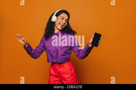 Happy woman with curly hair dances against a vibrant orange background, listening to music on her smartphone with headphones. She celebrates with colo Stock Photo