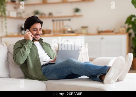 Man Talking on Phone, Working on Laptop in Living Room Stock Photo