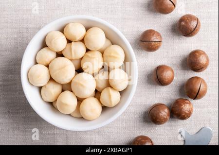 Roasted macadamia nuts in a white bowl on linen fabric. On the right fruits with sawn nutshells and opener key. Stock Photo