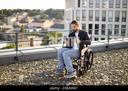 Man in wheelchair using phone and drinking coffee on rooftop Stock Photo