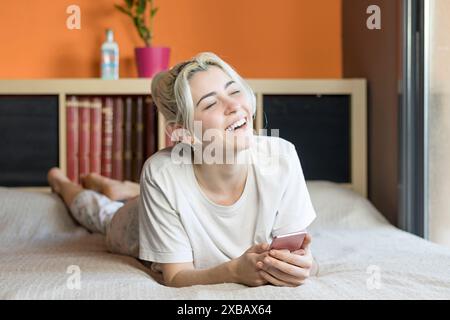 Blonde woman in a white shirt and floral pyjama bottoms lying on a bed, laughing while holding a phone. Background features an orange wall and bookshe Stock Photo