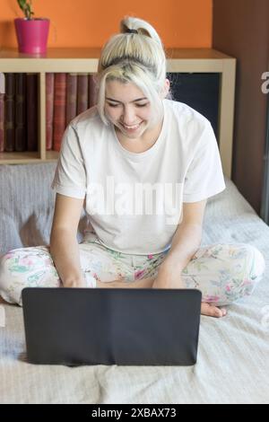Blonde woman with a ponytail, in a white t-shirt and floral pyjamas, sits cross-legged on bed, using a laptop. She is smiling, surrounded by books and Stock Photo