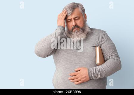 Portrait of sad senior man with book holding head on light blue background Stock Photo