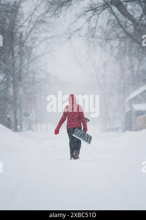 A woman in a red coat carries a snow shovel down a small-town road in the middle of a snowstorm Stock Photo