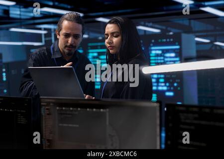 Programmers in server room checking recovery plan, monitoring tech gear energy consumption using notebook. Colleagues in data center workplace doing routine disk checks using laptop and tablet Stock Photo