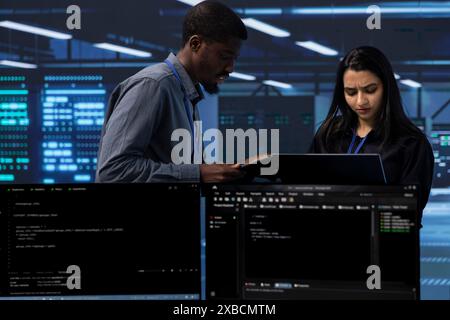 Engineers in server farm checking recovery plan, monitoring tech equipment energy consumption. Coworkers in data center workspace doing routine disk checks using laptop and tablet Stock Photo