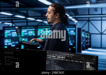 Engineer in data center using laptop to find firewall misconfigurations affecting rackmounts performance. Worker in server farm checking systems bottlenecks leading to sluggish data transfer rates Stock Photo