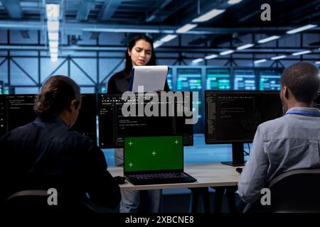 Manager inspecting server room equipment, reading paperwork from folder while engineers work on isolated screen laptop. Supervisor checking data center files next to chroma key device on desk Stock Photo