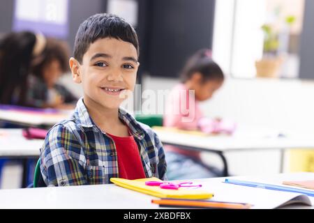 Biracial boy smiling at desk in classroom with school supplies around him. Other students and teacher in background focusing on their tasks and workin Stock Photo