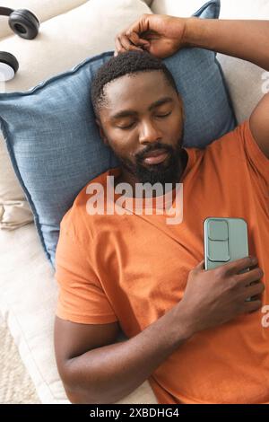 Young African American man relaxing on couch holding smartphone, at home. Cozy home environment with headphones nearby and soft pillows creating comfo Stock Photo