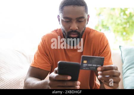An African American young man is holding credit card while using his smartphone, at home. He is sitting on a couch with a bright window and greenery i Stock Photo