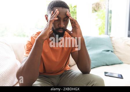 African American young man sits on couch, looking stressed with hands on temples. Bright living room with large windows and a smartphone on couch besi Stock Photo