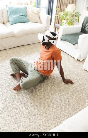 Young African American man wearing VR headset sitting on carpet. Living room with beige furniture and green plants in the background, creating a cozy Stock Photo