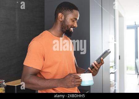 Young African American man holding tablet and a coffee mug, smiling at screen. He is standing in a modern kitchen with sunlight streaming through larg Stock Photo