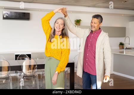 A biracial mature couple dancing together, smiling warmly at each other, at home. They are in modern, bright kitchen with clear chairs and a large din Stock Photo