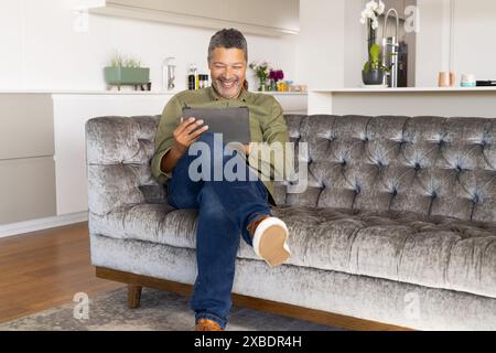 Mature biracial man sitting on couch, smiling while using a tablet, at home. Modern living room with a kitchen in background, featuring plants and sty Stock Photo