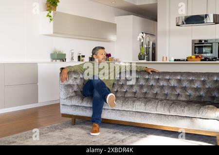 Mature biracial man sitting on couch in a modern living room, at home, copy space. Bright kitchen area with plants and appliances in white background, Stock Photo