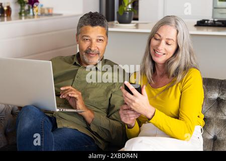 Biracial mature couple relaxing at home, interacting with laptop and smartphone, at home. Cozy living room with modern decor and kitchen in background Stock Photo