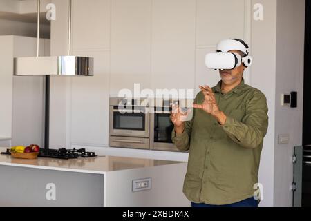 Mature biracial man wearing VR headset, interacting with virtual objects, at home, copy space. Modern kitchen with sleek appliances and a bowl of frui Stock Photo