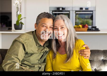 A biracial mature couple is smiling and sitting closely on couch at home, at home. They are in a modern kitchen with stainless steel appliances and a Stock Photo