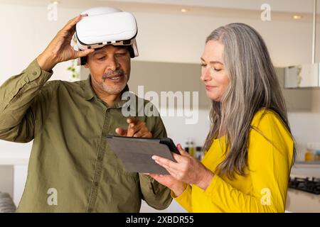A biracial mature couple using tablet and VR headset, at home. In modern kitchen with light-colored cabinets and countertops, engaging in a tech-relat Stock Photo
