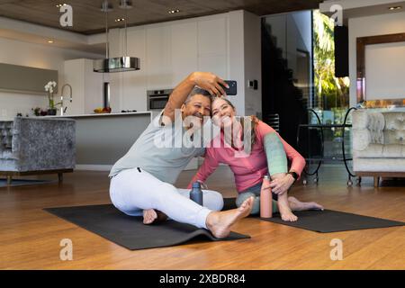 A biracial mature couple is sitting on yoga mats, smiling and taking selfie, at home. They are in a modern, spacious living room with wooden floors an Stock Photo