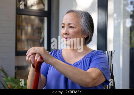 Asian mature woman holding cane, looking thoughtful on porch, at home. She is sitting with a modern home and garden in background, surrounded by green Stock Photo