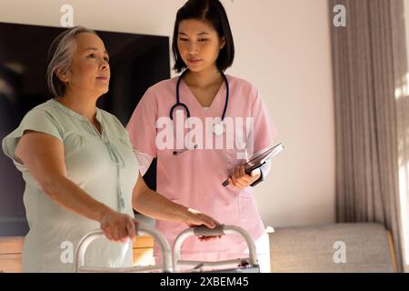 nurse healthcare worker helping older Asian woman use walker at home. They are in bright living room with a large TV and gray background curtains, una Stock Photo