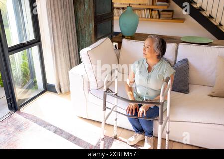 Asian senior woman using walker, sitting on couch in bright living room, at home Stock Photo