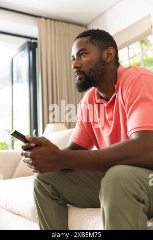 Young African American man holding remote control, watching tv, at home. Bright living room with large windows and beige background curtains, sunlight Stock Photo