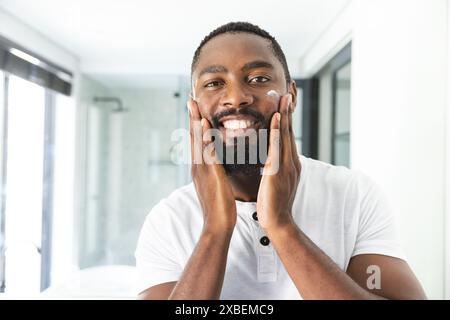 African American young man with beard smiling and touching his face, at home. Bright bathroom with glass shower and modern fixtures creating a clean, Stock Photo