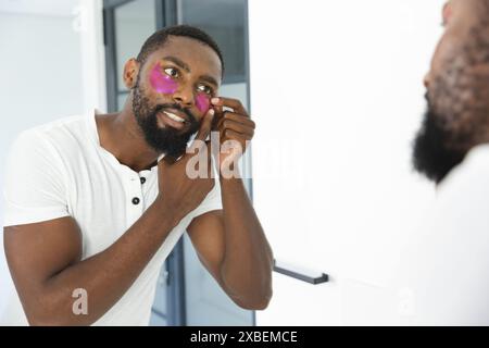 Young African American man applying under-eye patches while looking in mirror, at home. Modern bathroom with white walls and glass shower door in whit Stock Photo