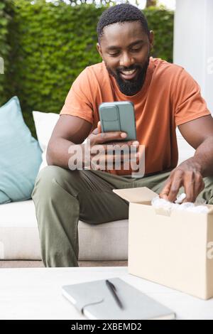 Young African American man smiling while using smartphone and opening package, at home. Outdoor patio with green foliage and comfortable seating in ba Stock Photo