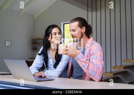 Young diverse couple are sitting together, using a laptop. They are in a modern home with large windows and minimalist decor, unaltered Stock Photo
