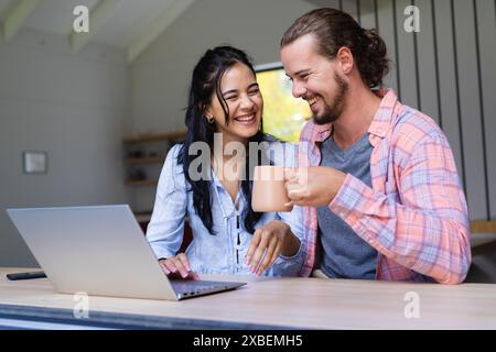 Young diverse couple are smiling while looking at a laptop. They are sitting at a kitchen counter with shelves and a window in background, unaltered Stock Photo