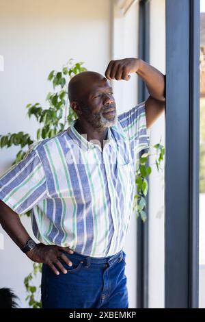 Mature African American man standing by window, looking thoughtful. Bright indoor space with large windows and green plants in background, creating se Stock Photo