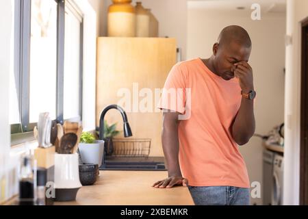 Young African American man in pink shirt standing stressed in modern kitchen. Bright natural light from large windows illuminates wooden cabinets and Stock Photo