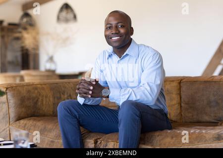 Young African American man sitting on brown couch, smiling confidently. He is in modern, well-lit living room with minimalist decor, featuring a white Stock Photo