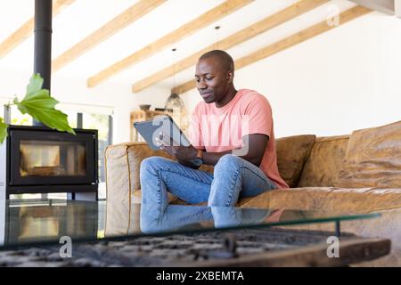African American young man sitting on couch using a tablet. Modern living room with wooden beams and a fireplace in background, creating a cozy and st Stock Photo