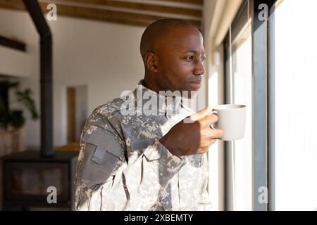 African American young soldier in military uniform holding mug and looking out window. Sunlight streaming through large windows, illuminating cozy, mo Stock Photo