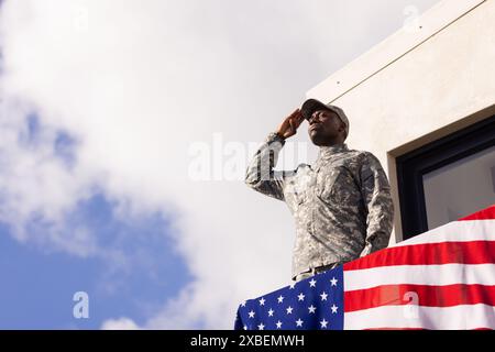 African American young soldier in military uniform saluting on balcony. American flag decorating balcony against a clear sky, creating a patriotic bac Stock Photo