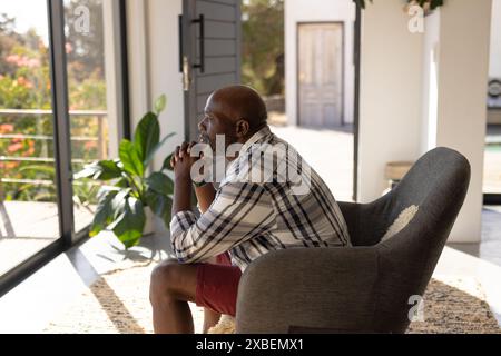 African American mature man sitting in chair, looking thoughtfully out window. Sunlight streams through large windows, illuminating a cozy, modern liv Stock Photo