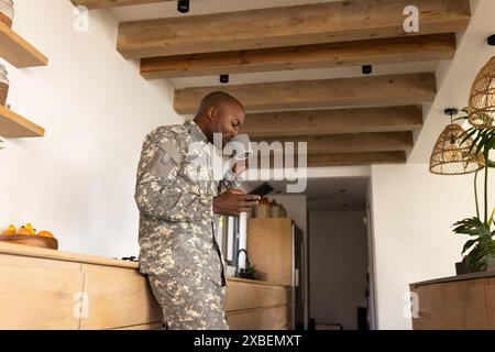 Young African American soldier in military uniform drinks coffee and checks smartphone. Modern kitchen with wooden beams and natural light creating wa Stock Photo