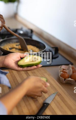 Young diverse couple preparing breakfast with avocados together in their kitchen. Modern kitchen with a wooden countertop, eggs, and a stove in backgr Stock Photo