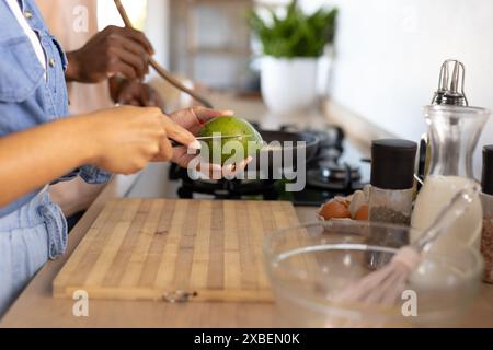 A young diverse couple are preparing breakfast with avocados in kitchen. They are standing at a wooden counter with various cooking ingredients and ut Stock Photo