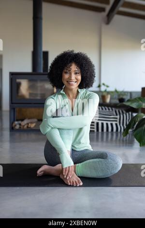 Young biracial woman sitting on yoga mat, looking relaxed and fit. Modern living room with fireplace and stylish decor in background, creating cozy an Stock Photo