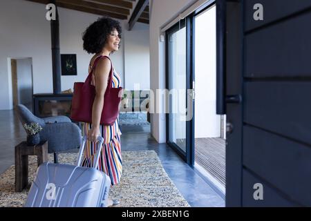 Biracial young woman with curly hair carrying suitcase and red bag, vacation home. Modern vacation home with large windows and stylish furniture, feat Stock Photo