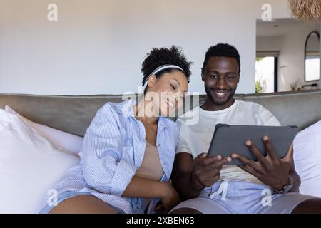 A diverse couple is relaxing on couch, smiling while looking at a tablet together. They are in a cozy living room with soft lighting and modern decor, Stock Photo